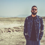 A man standing in desert with red sunglasses and a blue jean jacket