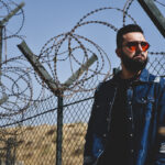 A man standing in desert with red sunglasses and a blue jean jacket