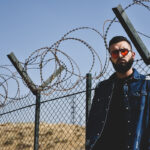 A man standing in desert with red sunglasses and a blue jean jacket